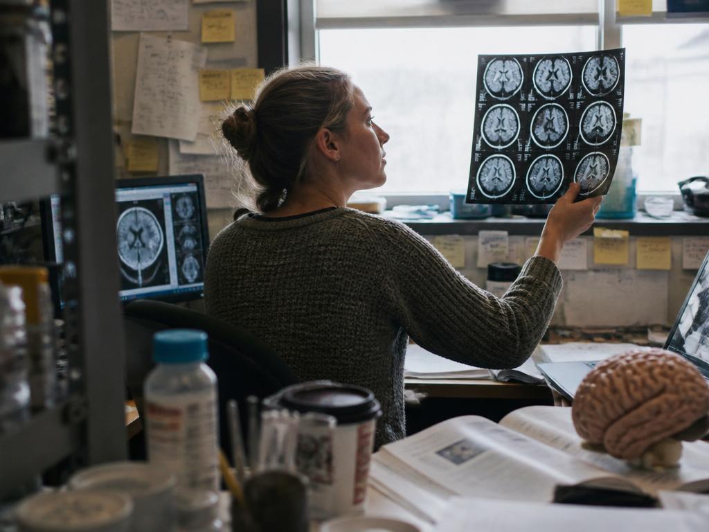 A neuroscience researcher holding a printed MRI brain scan up to the light at a cluttered lab desk