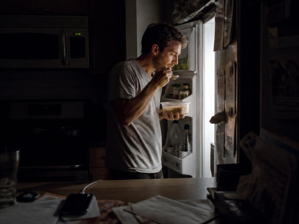 A man eating leftovers from a container at the open refrigerator late at night